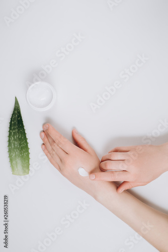 Top view woman putting nutritious cream on her hands on white background among jar of cosmetic cream, Aloe vera fresh leaves. Moisturizing cream for clean and soft skin. Healthcare concept.