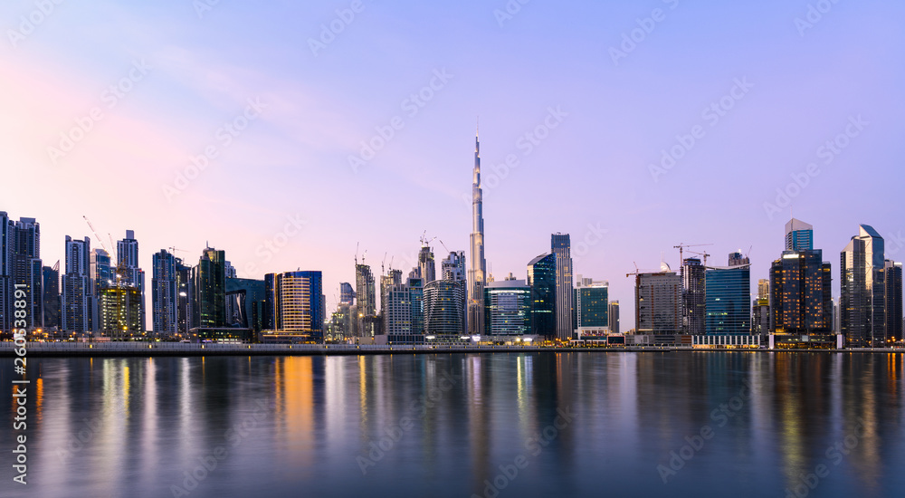 Panoramic view of the illuminated Dubai skyline during sunset with the magnificent Burj Khalifa and many others skyscrapers reflected on a silky smooth water flowing in the foreground. Dubai.