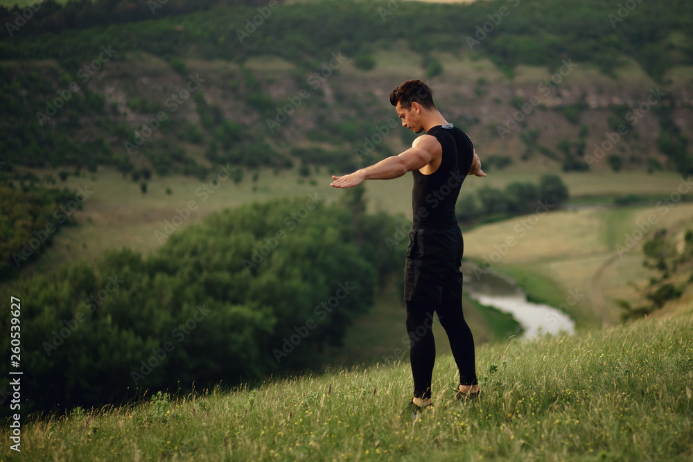 Athletic young man in sportswear doing exercises with raised hands and looking down on nature landscape background. Horizontal view. Athletic young man in sportswear doing exercises with raised hands and looking down on nature landscape background. Horizontal view.