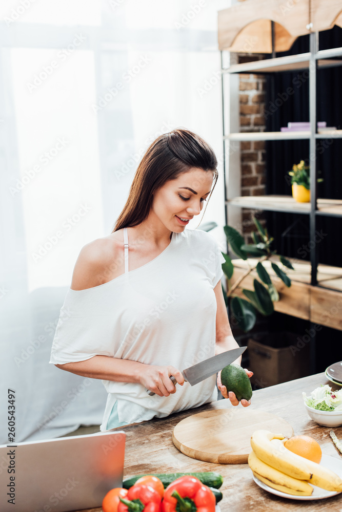 Smiling young woman cutting avocado at wooden table in kitchen Smiling young woman cutting avocado at wooden table in kitchen