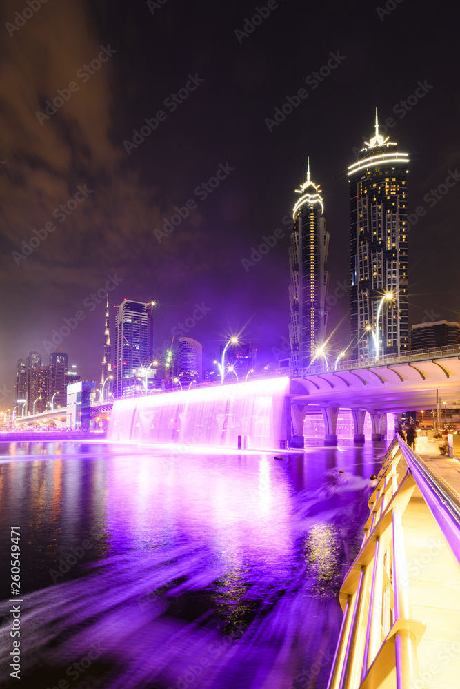Fototapeta premium Stunning view of the illuminated Dubai skyline with the magnificent Burj Khalifa in the background and the beautiful and colored waterfalls seen from the Dubai Water Canal boardwalk. Dubai.