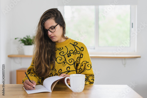 Mulher de óculos a escrever no seu caderno com uma caneca branca ao lado em cima da bancada de madeira.