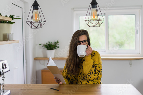 Mulher de óculos bebendo uma bebida numa caneca branca enquanto segura um caderno aberto na outra mão photo