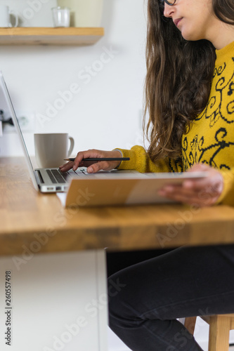 Mulher a trabalhar no seu computador portátil vista de lado. A segurar um caderno de apontamentos e com uma caneca branca do seu lado direito.