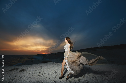 Beautiful brunette model girl with long sexy legs in fashionable lace dress posing at the sea coast at the sunset with amazing beautiful sky