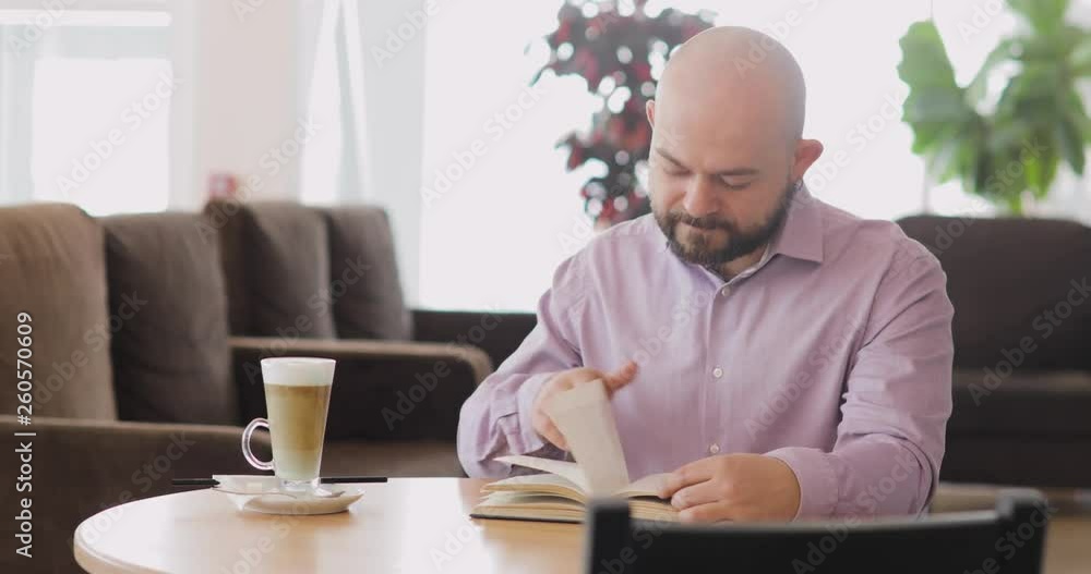 Bearded bald man is reading a book sitting at table in hotel hall cafe with cup of cappuccino. Front view. Bearded bald man is reading a book sitting at table in hotel hall cafe with cup of cappuccino. Front view.