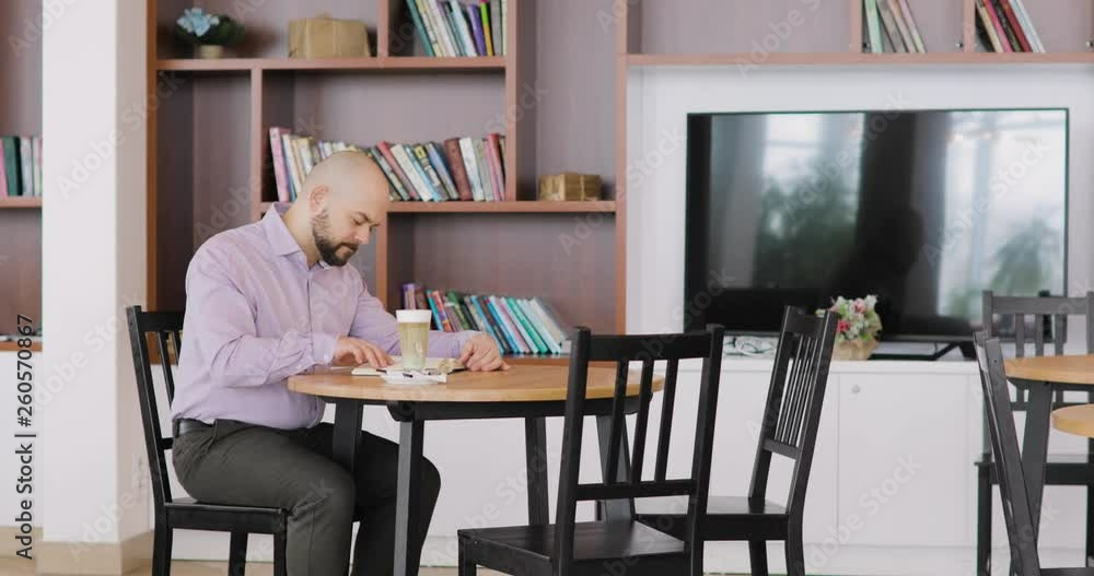 Bearded bald man in office clothes is reading a book sitting in hotel hall cafe with cup of cappuccino. Side view. Bearded bald man in office clothes is reading a book sitting in hotel hall cafe with cup of cappuccino. Side view.