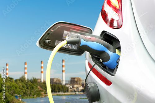 plug of power cable electric supply during charging at ev car (electric vehicle charging) on electric power plant with smokestack and blue sky background