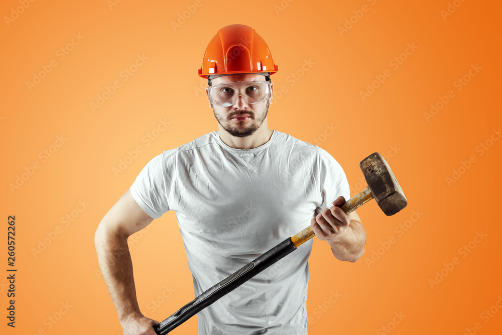 Bearded Man in a helmet holds a sledgehammer on an orange background. Concept of construction, contractor, repair. Bearded Man in a helmet holds a sledgehammer on an orange background. Concept of construction, contractor, repair.