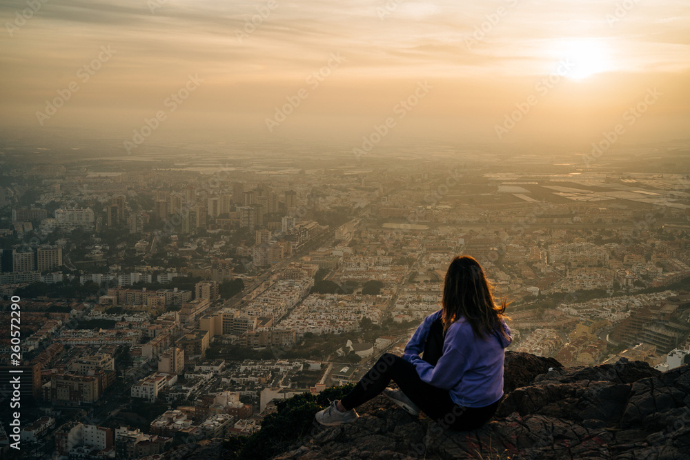 Young woman sitting and using the mobile on the top of the mountain looking at the city from above, during nice sunset Young woman sitting and using the mobile on the top of the mountain looking at the city from above, during nice sunset