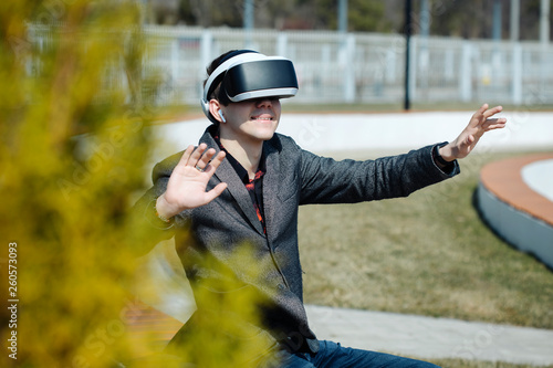Young modern man wearing virtual reality glasses in a city park sits in a park on a bench and manipulates his hands, playing with reality