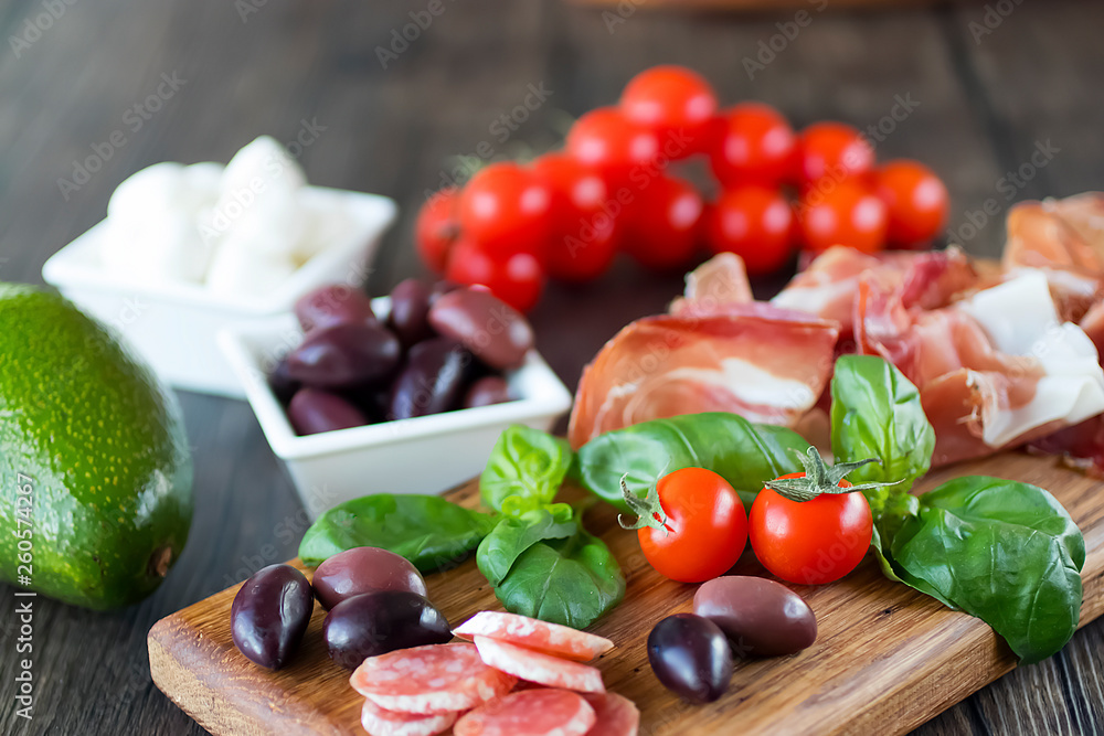 Prosciutto, bread, olives, walnut, mozzarella, salami, basil and cherry tomatoes on  brown wooden board.  Mediterranean kitchen.