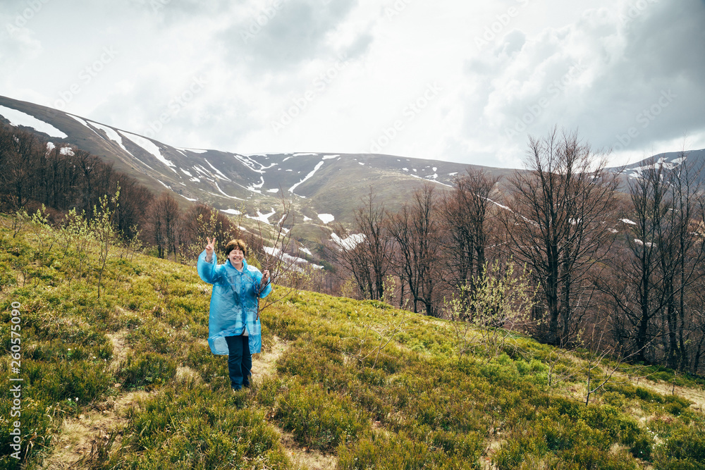 Happy smiling traveller senior beautiful woman in blue rain jacket and jeans in mountains surrounded by forest, enjoying silence and harmony of nature