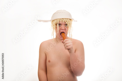 A young, fat, cheerful, smiling boy eating sausage and having a colander and spaghetti on his head. Plump body without clothing. White background. Portrait photo.