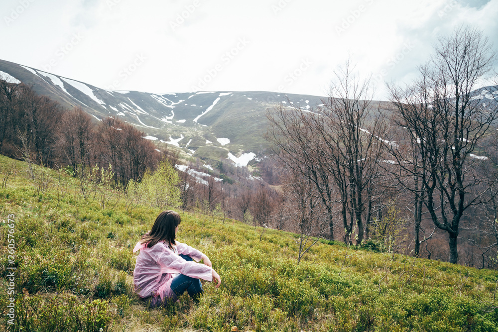 Tourist girl in pink rain jacket sitting on the grass looking at mountains surrounded by forest, enjoying silence and harmony of nature