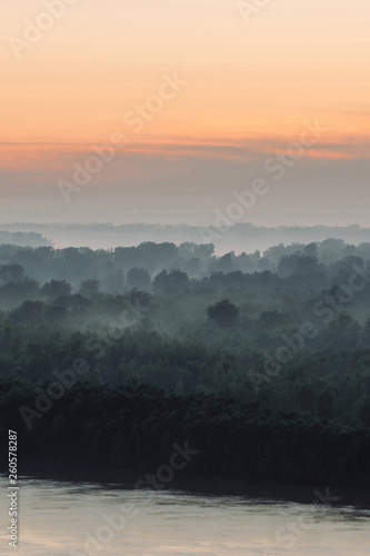 Mystical view on riverbank of large island with forest under haze at early morning. Eerie mist among layers from tree silhouettes under predawn sky. Morning atmospheric landscape of majestic nature.
