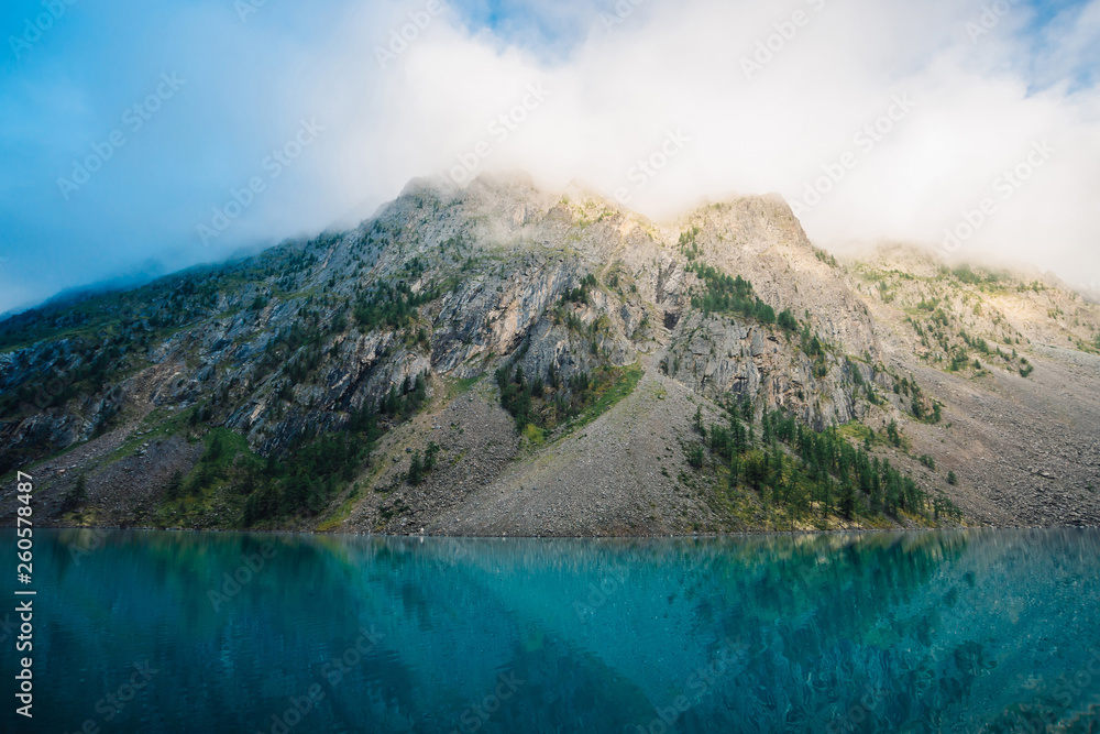 Giant cloud above rocky ridge with trees in sunlight. Amazing mountain lake. Mountain range under blue cloudy dawn sky. Wonderful rocks. Morning landscape of highland nature. Low clouds in sunny light
