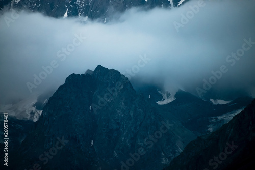 Low cloud before huge glacier. Giant snowy rocky mountain wall in thick fog. Early morning in mountains. Impenetrable fog. Dark atmospheric foggy landscape with cold rocks. Tranquil mystic atmosphere.
