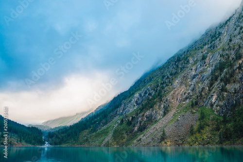 Giant cloud above mountainside with trees in sunlight. Amazing mountain lake. Mountain range under blue cloudy dawn sky. Wonderful rocks. Morning landscape of highland nature. Low cloud in sunny light