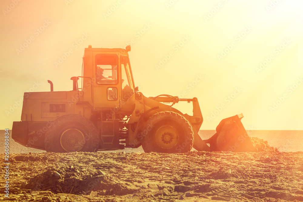 Yellow bulldozer on the background of sunset sky and seashore. Copy space and light from the right side. Tint Yellow bulldozer on the background of sunset sky and seashore. Copy space and light from the right side. Tint