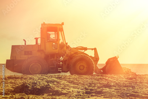 Yellow bulldozer on the background of sunset sky and seashore. Copy space and light from the right side. Tint