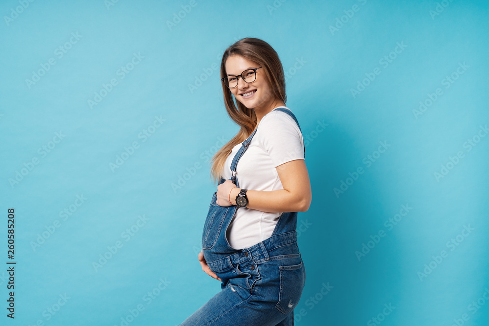 Smiling pregnant woman wearing glasses caressing her belly over blue background Smiling pregnant woman wearing glasses caressing her belly over blue background