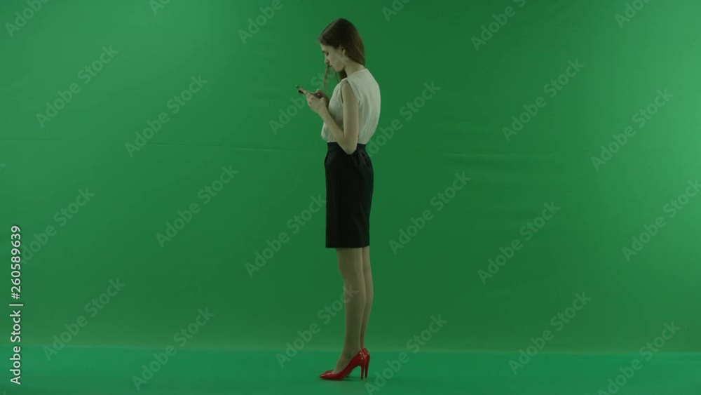 beautiful young business woman in white shirt and black skirt is using her mobile phone standing sideways against green screen. beautiful young business woman in white shirt and black skirt is using her mobile phone standing sideways against green screen.
