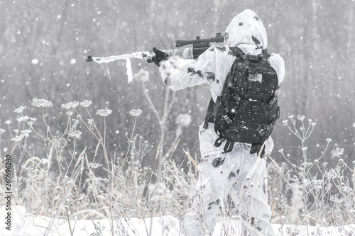 Man soldier in the winter on a hunt with a sniper rifle in white winter camouflage aiming standing in the snow