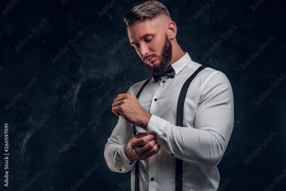 Stylishly dressed young man in shirt with bow tie and suspenders. Studio photo against dark wall background
