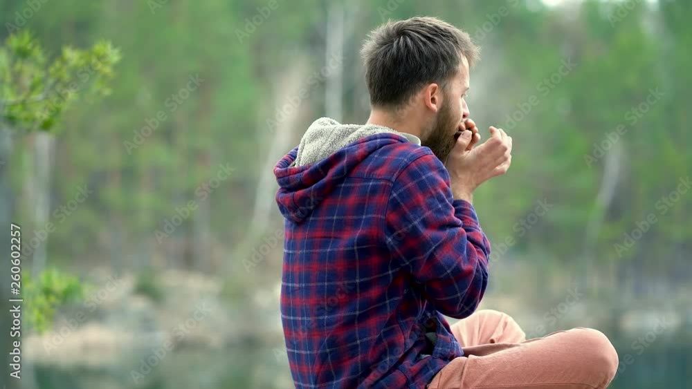 Young beard man playing on the harmonica. Sitting on the rock near the lake. Steadycam shot. Young beard man playing on the harmonica. Sitting on the rock near the lake. Steadycam shot.