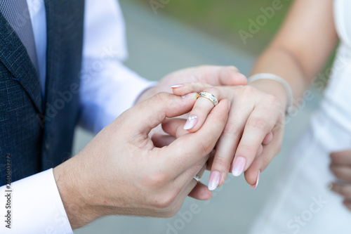 A man in a vest and tie wearing a gold wedding ring to a woman in a wedding dress on the ring finger.