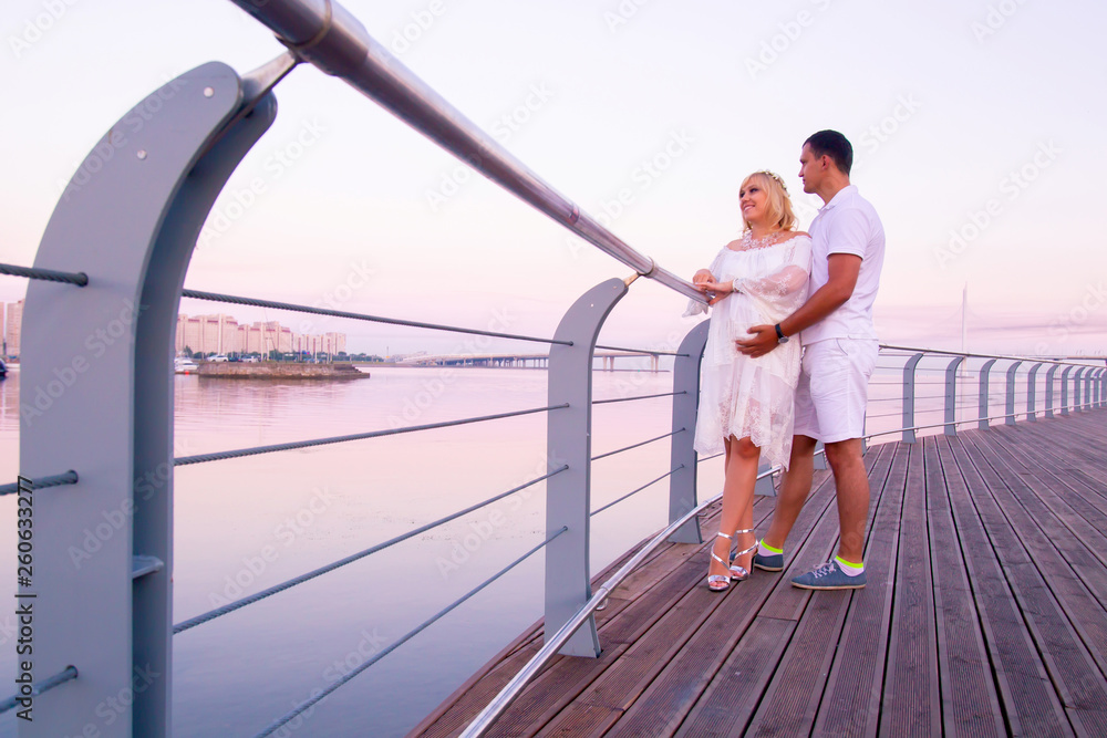 A married couple is expecting a child. Pregnancy. A man hugs a pregnant woman. Man and woman on the waterfront. Married couple in white clothes.