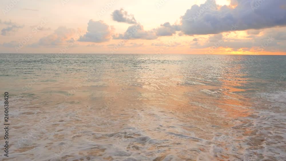 Waves of warm turquoise water smash against the sand of an island beach as a colorful sunset lights the sky on the horizon. Dramatic evening paradise scenery as the sun sets on an empty beach. Waves of warm turquoise water smash against the sand of an island beach as a colorful sunset lights the sky on the horizon. Dramatic evening paradise scenery as the sun sets on an empty beach.