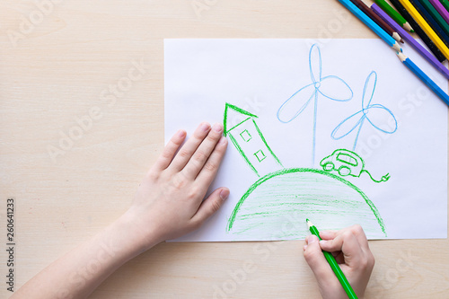 The child draws with colored pencils a green house, an electric car and a windy power station. Concept of ecology