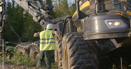 The operator of the logging machine to cutting trees in white helmet and green jumpsuit went into the forest to inspect equipment after work
