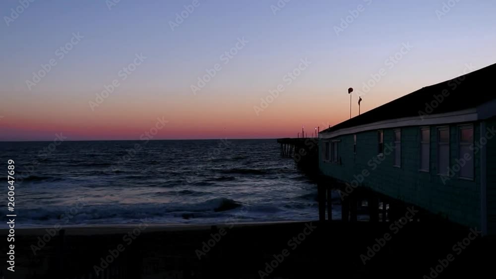 Clear sky at sunrise in the Outer Banks of North Carolina as waves roll onto the sand beside a wooden fishing pier. Gradient morning sky with no clouds. Clear sky at sunrise in the Outer Banks of North Carolina as waves roll onto the sand beside a wooden fishing pier. Gradient morning sky with no clouds.
