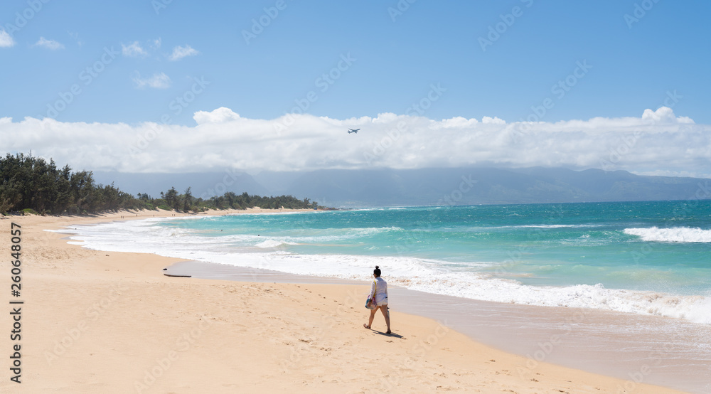 Asian woman walking across the tropical sandy beach with turquoise waves crashing on it. Plane flies away in the background.