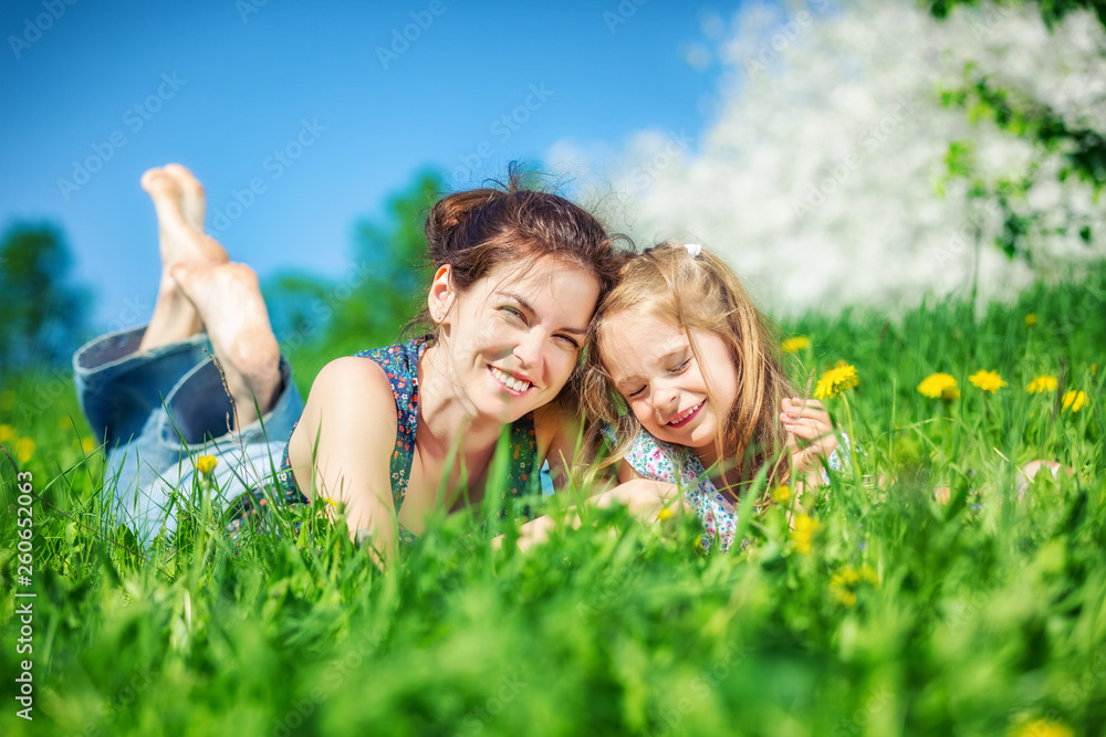Young woman and her daughter lying on green summer grass with blooming yellow dandellions Young woman and her daughter lying on green summer grass with blooming yellow dandellions