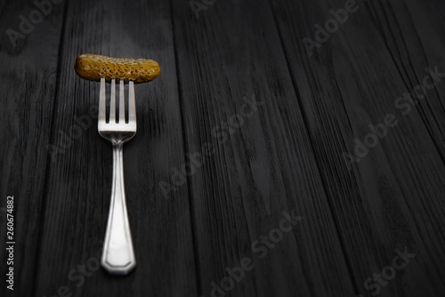 Fork with pickled cucumer on black wooden table background.