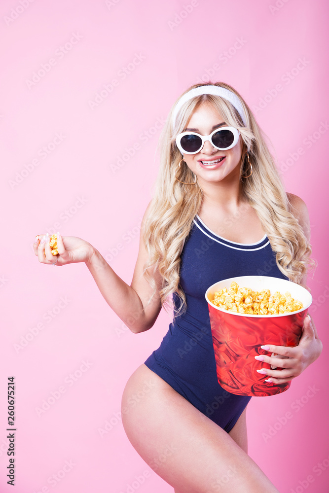 Obraz premium Amazing cute young pretty girl in sunglasses on the pink background eats popcorn and looking at the camera, wearing a blue swimsuite.