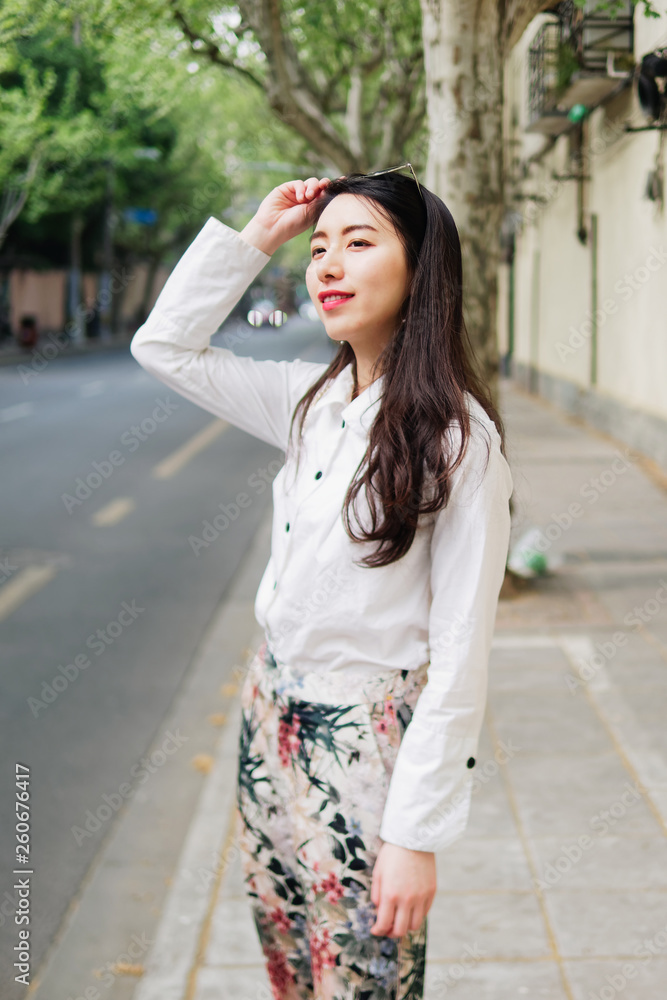 Outdoor portrait of young beautiful fashionable Chinese lady in casual clothes standing and smiling looking away in sunny Shanghai street. Female fashion. City lifestyle concept.