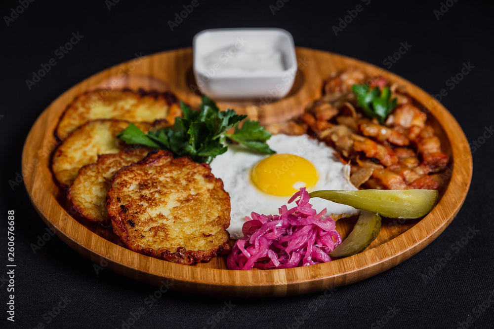Potato pancakes with a fried egg, bacon, sourcream, horse radish, pickles and parsley on a wooden plate on black background Potato pancakes with a fried egg, bacon, sourcream, horse radish, pickles and parsley on a wooden plate on black background