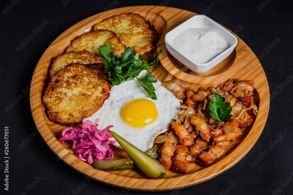 Potato pancakes with a fried egg, bacon, sourcream, horse radish, pickles and parsley on a wooden plate on black background Potato pancakes with a fried egg, bacon, sourcream, horse radish, pickles and parsley on a wooden plate on black background