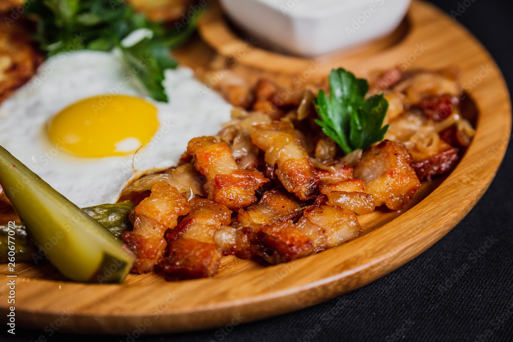 Potato pancakes with a fried egg, bacon, sourcream, horse radish, pickles and parsley on a wooden plate on black background Potato pancakes with a fried egg, bacon, sourcream, horse radish, pickles and parsley on a wooden plate on black background