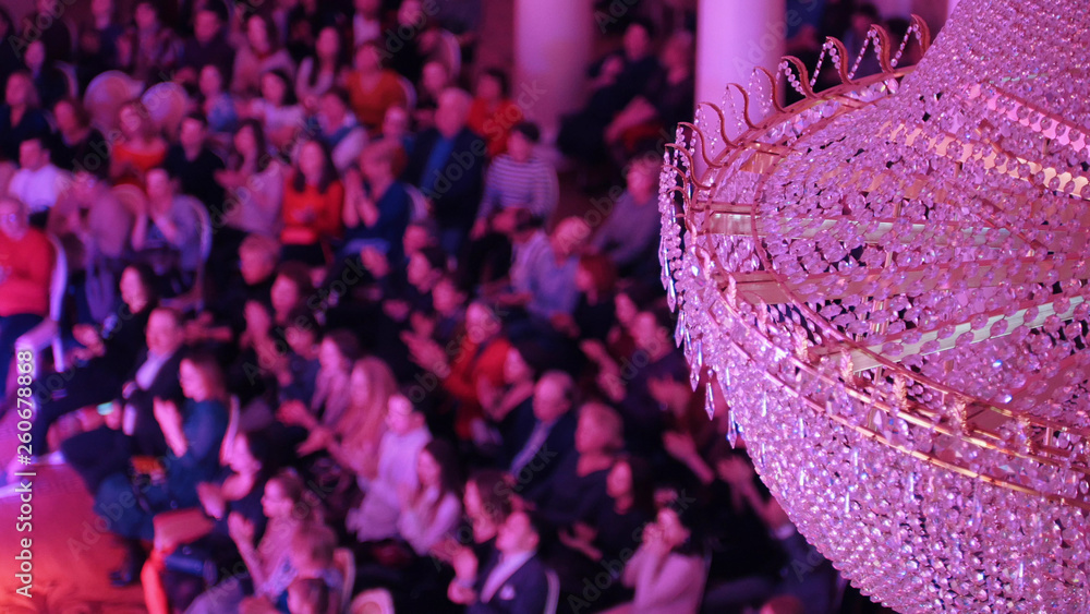 Obraz premium A concert in the concert hall with a big crystal chandelier. People sitting on the chairs and clapping their hands