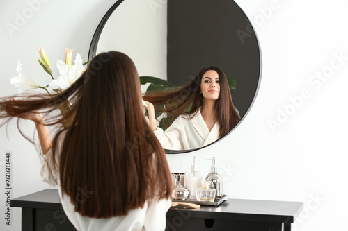 Beautiful young woman with healthy long hair looking in mirror at home