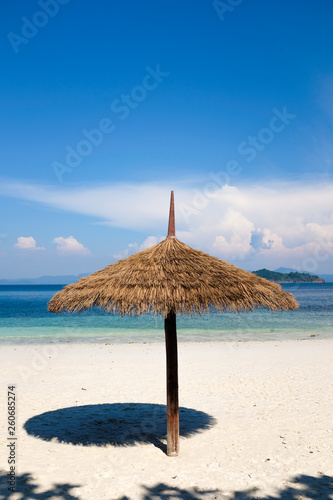 Natural palm leaves umbrella on white sand beach and bright blue sea water in background under crystal clear bright blue sky in sunny day. Tropical summer season vacation travel.