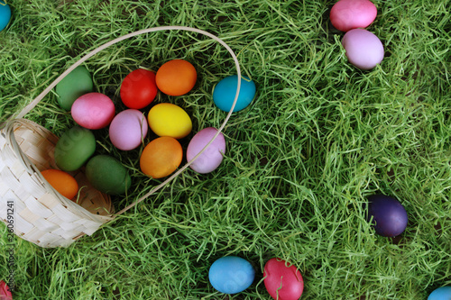 Colorful eggs scattered from basket on green grass. Easter background or egg hunt concept. Pink, yellow, green, orange eggs, hay, top view