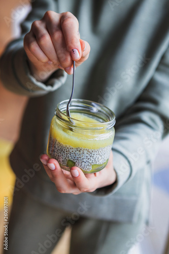 An attractive girl, a brunette, holds a jar with a sweet dessert of kiwi, chia and pineapple in her hand, enjoying a meal in the interior of a cozy cafe near the panoramic window with morning light