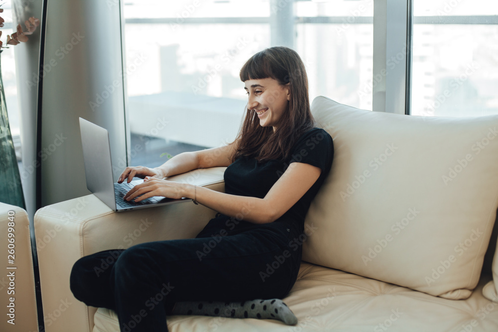 Happy smiling hipster girl typing on a laptop computer keyboard, chatting with family or friends via online messenger while sitting on a cozy sofa ot home with modern interior design Happy smiling hipster girl typing on a laptop computer keyboard, chatting with family or friends via online messenger while sitting on a cozy sofa ot home with modern interior design